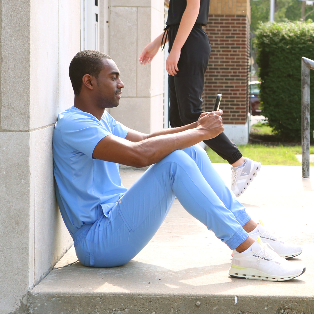 Man Texting on Phone Sitting in Ceil Blue Men&#39;s Scrub Joggers and Double-Pocket Scrub Top